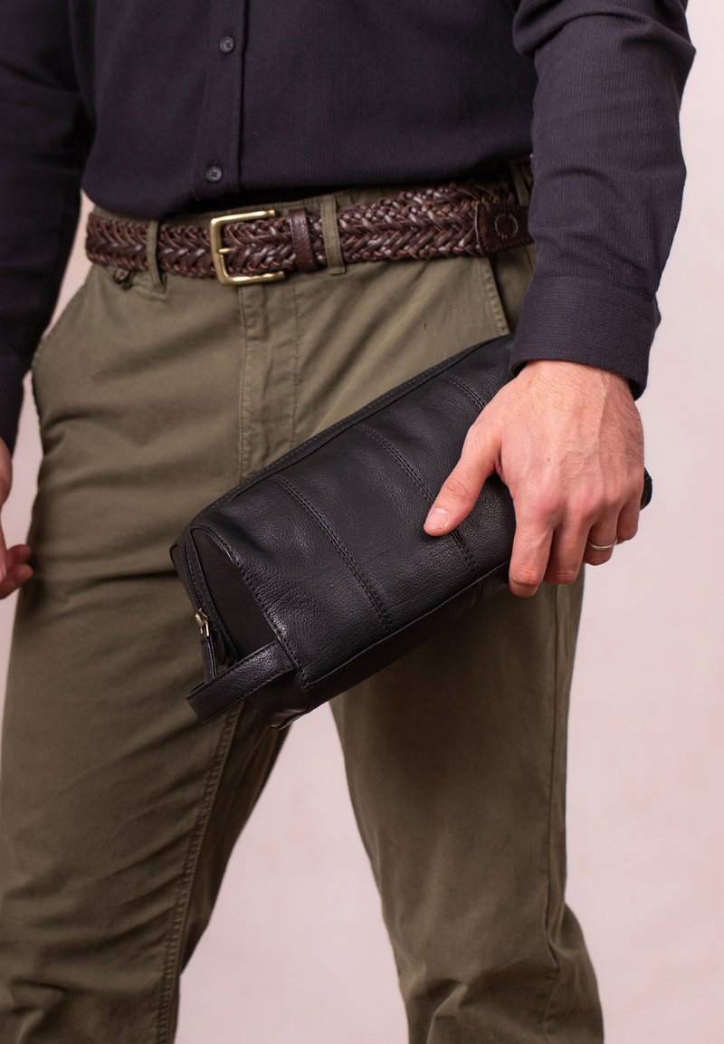 Man in olive pants and black shirt holding a black leather clutch bag with a zipper and wrist strap by his side.