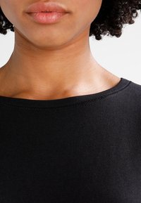 Detailní záběr na dolní část obličeje a krk osoby se středně hnědou pokožkou, přirozenými rty, kudrnatými vlasy, která nosí černé tričko s kulatým výstřihem.