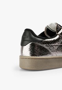 Zapatilla plateada metálica con parte superior texturizada, talón de cuero negro y suela de goma beige. Presenta un acabado suave y materiales en contraste.