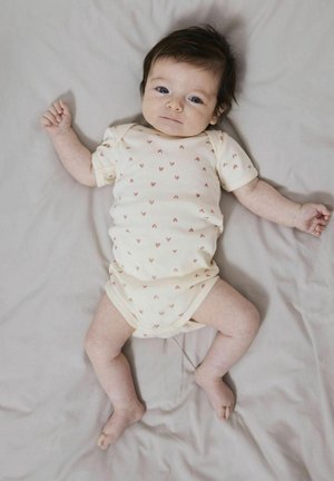 Vaaleanbeige vauvan body, jossa on pieniä sydänkuvioita, lyhyet hihat ja napsautettavat sulkimet. Vauva makaa pehmeällä, neutraalivärisellä huovalla.
