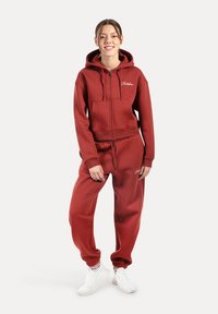 Bordowy hoodie z przednimi kieszeniami i zamkiem, zestawiony z pasującymi dresami. Białe logo i elastyczne mankiety. Białe sneakersy.