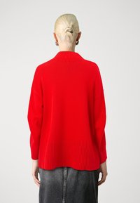 Pull en maille rouge avec un col et un ourlet côtelés, présentant une coupe ample et des manches longues. Texture lisse et couleur unie sans motifs.