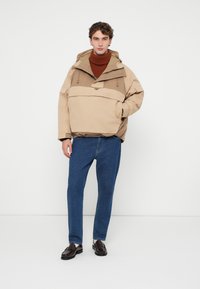 Veste à capuche beige avec une poche frontale, associée à un jean bleu et des mocassins foncés. La veste présente une coupe décontractée et des panneaux contrastants.