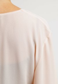 Gros plan du haut du dos et de l’épaule d’une personne portant un haut en tissu beige clair texturé avec un col rond.