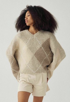 Kvinde med krøllet hår iført en beige og brun argyle trøje og lysebeige shorts, der kigger til siden mod en ensfarvet baggrund.