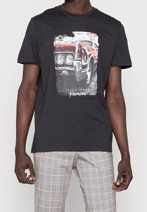 Mężczyzna w czarnej koszulce z grafiką vintage czerwonego samochodu z przodu i napisem „Jack & Jones Premium” oraz w spodniach w kratę.