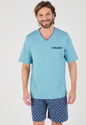 Uomo sorridente che indossa una maglietta azzurro chiaro a maniche corte con scollo a V e tasca, e pantaloncini blu navy con fantasia di ancore rosse e bianche.