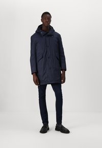 Navy waterproof parka with a hood, front button closure, two exterior pockets, and sleeves with elastic cuffs. Worn over dark jeans and black shoes.