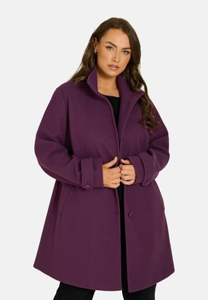 Donna che indossa un cappotto viola con bottoni e ampie maniche a risvolto, in piedi e che sistema la parte davanti su uno sfondo chiaro e uniforme.