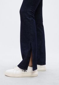 Σκούρα μπλε τζιν με καμπάνα και πλευρικές σχισμές στο κάτω μέρος, συνδυασμένα με λευκά αθλητικά παπούτσια που έχουν υφή στη σόλα και διακριτικό λογότυπο.