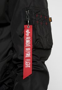 Schwarze Stoffjacke mit einer Reißverschlusstasche und einem auffälligen roten Etikett mit weißem Text, das "REMOVE BEFORE FLIGHT" (VOR DEM FLUG ENTFERNEN) liest. Strukturierter Stoff.