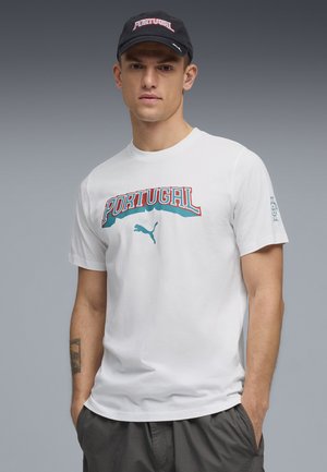 Homme portant une casquette noire « Portugal » et un t-shirt blanc avec le texte « Portugal » et le logo Puma, debout les mains dans les poches sur fond gris.