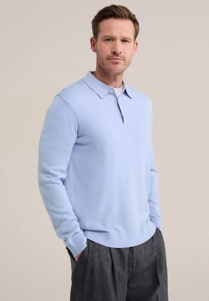 Homme aux cheveux châtain clair porte un pull polo bleu clair à manches longues et un pantalon gris foncé, debout les mains dans les poches devant un fond neutre.