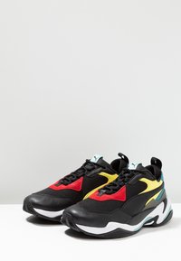 Czarne sportowe buty z czerwonymi, żółtymi i turkusowymi akcentami, wykonane z siateczki i zamszu, o teksturowanej powierzchni i grubej gumowej podeszwie.