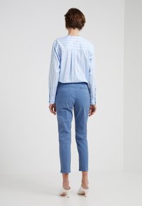 Personne se tenant de dos, portant une chemise rayée bleu clair, un pantalon bleu et des chaussures à talons blancs devant un fond léger uniforme.