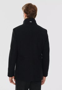 Czarny płaszcz z wełną o wysokim kołnierzu, z guziczkami na plecach i dopasowanym kroju. Gładka tekstura i czyste linie tworzą wyrazisty design.