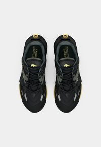 Black and gray Lacoste sneakers with yellow accents viewed from above, showing laces, logo, and cushioned interior.