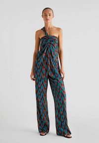 Ein Ein-Schulter-Jumpsuit in Teal, Orange und braunem Ikat-Muster. Hergestellt aus leichtem Stoff mit einem geknoteten Oberteil und weitem Bein.