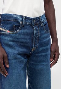 Gros plan d’une personne portant un jean en denim bleu avec des passants de ceinture visibles, un bouton avant, une braguette à fermeture éclair et une petite étiquette sur la poche pour la monnaie.