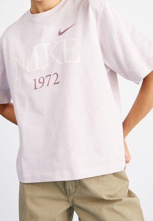 Osoba w jasnoróżowej koszulce Nike z napisem i logo "NIKE 1972", w połączeniu z beżowymi spodniami, ręce spoczywają na biodrach.