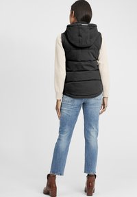 Svart vadderad väst med huva, kombinerad med en krämfärgad långärmad skjorta. Bärs med ljusblå raka jeans och bruna ankelstövlar.