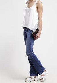 Vit ärmlös linne, blå utsvängda jeans, vita plattformssandaler, håller en liten svart och röd clutch. Klara linjer och avslappnad passform.