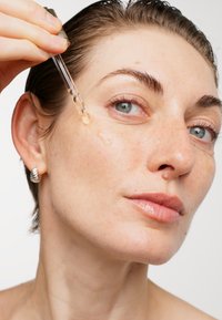 Siero per il viso trasparente applicato con un contagocce. Il modello ha capelli corti e bagnati, pelle liscia e indossa un orecchino a cerchio d'argento.