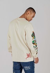 Beige Langarmhemd mit buntem Totenschädel und Design auf dem linken Ärmel; aus Baumwollmaterial mit rotem Logo-Detail am Saum.