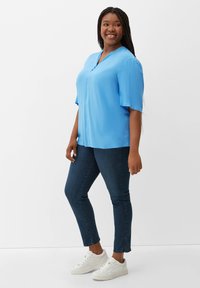 Light blue short-sleeve blouse with a V-neck and three buttons, paired with dark denim jeans and white sneakers, standing on a plain background.