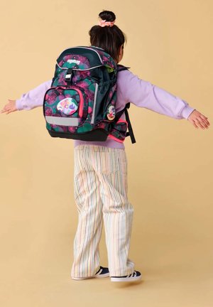 Kind trägt gestreifte Hose und lila Sweatshirt mit großem buntem Rucksack, Arme ausgestreckt, steht vor beigem Hintergrund.