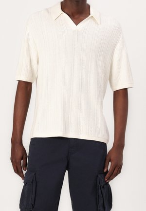 Polo a maniche corte color crema in maglia, con scollo a V, caratterizzato da una trama a coste verticali e una vestibilità rilassata. Abbinata a pantaloni cargo scuri.