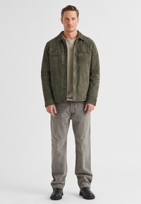 Wildlederjacke in Olivgrün mit zwei Brusttaschen, Knopfverschluss und kragenlosem Design, kombiniert mit hellgrauen Jeans und schwarzen Stiefeln.