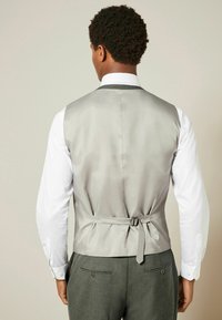 Chaleco gris de satén para hombre, con un respaldo entallado y un detalle de lazo, llevado sobre una camisa blanca y combinado con pantalones gris oscuro.