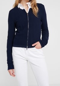 Cárdigan de cremallera azul marino con textura de canalé, diseño ajustado y mangas largas, combinado con pantalones blancos. Detalle de cremallera plateada en la parte frontal.