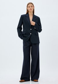 Blazer azul marino con grandes botones plateados, dos bolsillos frontales y un corte a medida, combinado con pantalones de pinstripe azul marino de pierna ancha. Zapatos marrones.