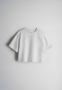 T-shirt court gris clair à manches courtes suspendu contre un mur blanc uni, vue de face avec encolure ronde.