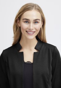 Melna jaka ar augstu apkakli un bez aizdare, kas uzlikta uz tumša tops ar mežģīņu akcentu. Gluda auduma ar matētu apdari.