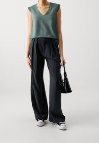 Grön ärmlös topp med spetsdetaljer, matchad med lösa svarta byxor. Modellen håller en liten svart handväska och bär svarta och vita sneakers.