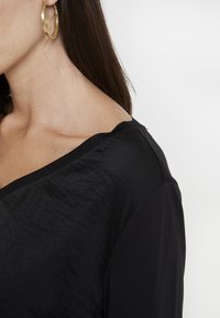 Černý top s texturovaným, plynulým designem a výstřihem na ramena, doplněný kontrastním hladkým lemováním po okrajích.