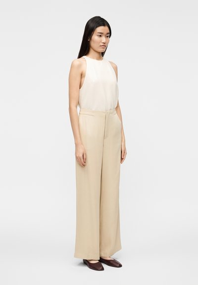 Smėlio spalvos be rankovių palaidinė, laisvai krentančios kreminės kelnės, elegantiški bordo spalvos batai. Lygus audinys su subtilia danga, tiesi silueto linija, minimalus dizainas.