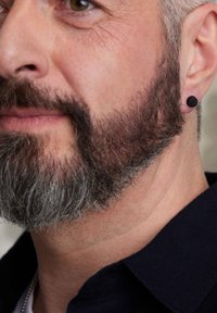 Black stud earring on the left ear of a man with a short, gray beard. He has olive skin, wearing a dark polo shirt. Close-up view.