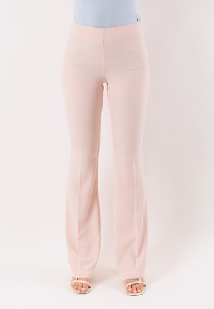 Donna che indossa pantaloni a vita alta color rosa chiaro a vita alta e svasati, con sandali beige aperti, in piedi davanti a uno sfondo bianco.