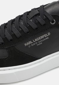 Svart lær- og semsket skinnsneaker med teksturerte svarte lisser, hvit gummisåle og preget logo-detalj på siden.