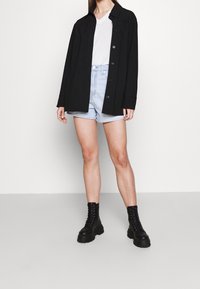 Giacca nera con bottoni sopra una maglietta bianca a scollo a V, abbinata a pantaloni corti di denim azzurri e stivali alla caviglia neri con lacci. Texture liscia.
