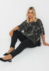 Top trasparente nero con motivo floreale bianco, abbinato a pantaloni neri slim-fit e tacchi slingback neri, caratterizzato da una vestibilità rilassata e fessure laterali.