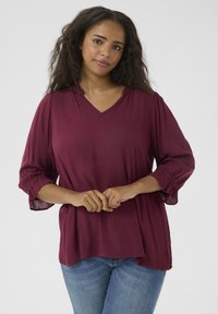Blouse bordeaux avec un col en V, manches trois-quarts avec des poignets élastiques, tissu léger et coupe décontractée, associée à un jean bleu.