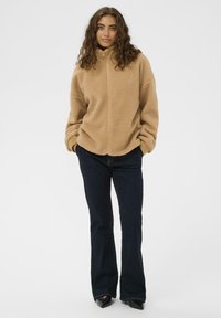 Beež fliiskindaga jope kõrge kaelusega, lukuga ja pikkade varrukatega, kombineerituna tumehallide flöödidest teksade ja mustade kingadega.