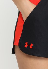 Pantalones cortos deportivos negros con detalles rojos, con una textura suave, dobladillo dividido y un logotipo visible en el lado inferior izquierdo.