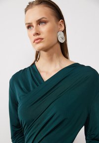 Teal draped top in smooth fabric with a V-neckline and gathered side details, paired with large white earrings featuring a marbled design.
