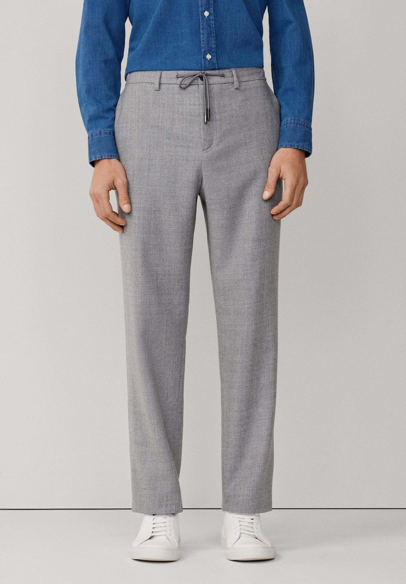 Uomo che indossa pantaloni su misura grigio chiaro con vita regolabile, scarpe da ginnastica bianche e una camicia di jeans blu abbottonata.